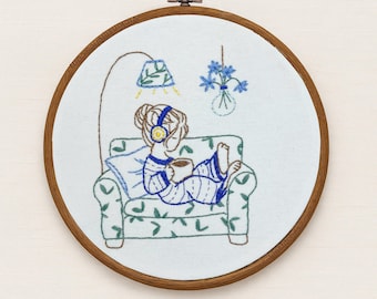 May include: Embroidered hoop art featuring a person relaxing on a floral couch, wearing headphones, and holding a mug. The scene includes a lamp and flowers, all in a cozy, hand-stitched style. The wooden hoop is approximately 8 inches in diameter.