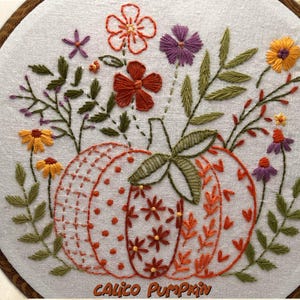 May include: Embroidered artwork featuring a pumpkin design with floral accents. The pumpkin is orange and white, with various stitch patterns. Surrounding the pumpkin are embroidered flowers in orange, purple, and yellow, with green leaves. The text "Calico Pumpkin" is at the bottom.