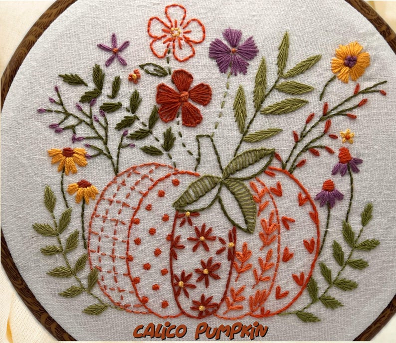 May include: Embroidered artwork featuring a pumpkin design with floral accents. The pumpkin is orange and white, with various stitch patterns. Surrounding the pumpkin are embroidered flowers in orange, purple, and yellow, with green leaves. The text "Calico Pumpkin" is at the bottom.
