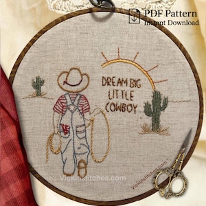 May include: Embroidered hoop art with a cowboy theme. The design shows a cowboy in dungarees, a lasso, cacti, and the text "Dream Big Little Cowboy." The embroidery is on a beige fabric, set in a wooden hoop.