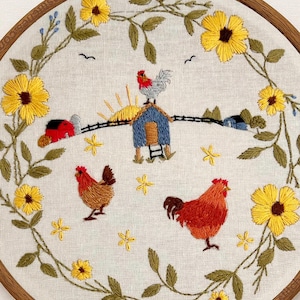 May include: A circular embroidery hoop featuring a farm scene with two chickens, a blue coop, a red barn, and a rising sun. The scene is framed by a wreath of yellow sunflowers and green leaves on a light beige fabric background.