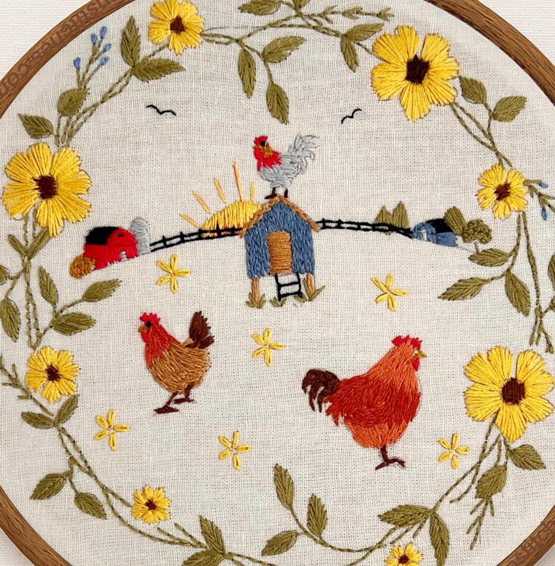 May include: A circular embroidery hoop featuring a farm scene with two chickens, a blue coop, a red barn, and a rising sun. The scene is framed by a wreath of yellow sunflowers and green leaves on a light beige fabric background.