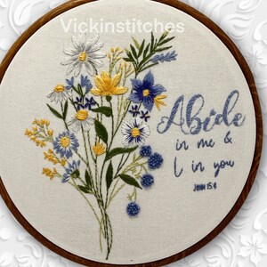 May include: Embroidered floral hoop art featuring a bouquet of white, yellow, and blue flowers. The text "Abide in me & I in you" is stitched in blue thread, with "John 15:4" below. The artwork is set in a wooden hoop.