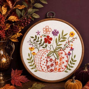 May include: A circular wooden embroidery hoop containing white fabric with a hand-stitched design of a pumpkin decorated with various red and orange floral patterns, surrounded by embroidered green leaves, yellow daisies, and purple wildflowers.