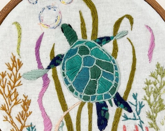 May include: A circular wooden embroidery hoop containing a white fabric panel with a hand-stitched sea turtle in shades of teal, green, and blue. The scene includes embroidered seaweed in yellow, pink, and green, with three small bubbles above.