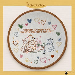May include: A circular wooden embroidery hoop containing white fabric with hand-stitched illustrations of Winnie the Pooh, Piglet, Eeyore, and Rabbit sitting on a log. The text reads, "Sometimes the smallest things take up the most room in your heart," surrounded by colorful embroidered hearts.