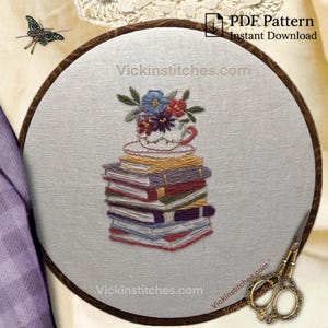 May include: Embroidered artwork featuring a stack of books topped with a teacup overflowing with flowers. The design is set within a wooden embroidery hoop. The text "Vickinstitches.com" is visible. The words "PDF Pattern Instant Download" are also present.