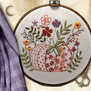 May include: Embroidered pumpkin with floral accents in a wooden hoop. The pumpkin is orange and white with a dotted and heart pattern. The flowers are red, orange, and purple. The embroidery is on a white fabric.