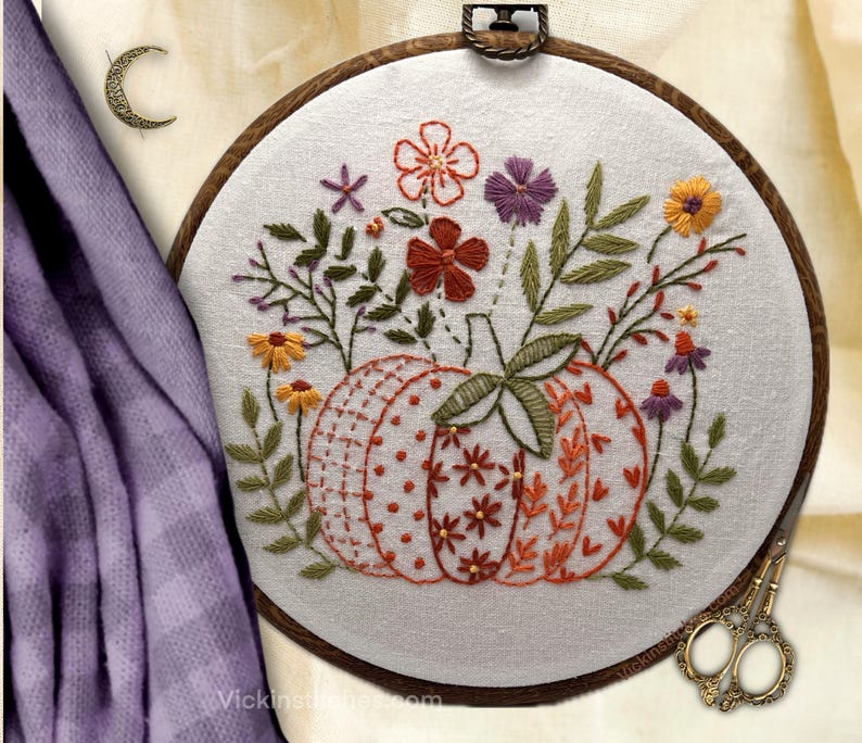 May include: Embroidered pumpkin with floral accents in a wooden hoop. The pumpkin is orange and white with a dotted and heart pattern. The flowers are red, orange, and purple. The embroidery is on a white fabric.