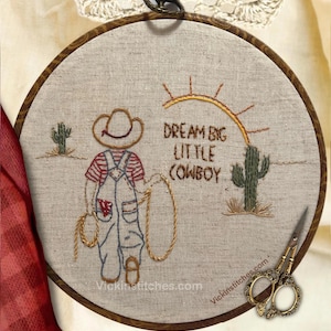 May include: Embroidered hoop art featuring a cowboy with a lasso, two cacti, and a sun. The text reads "Dream Big Little Cowboy".