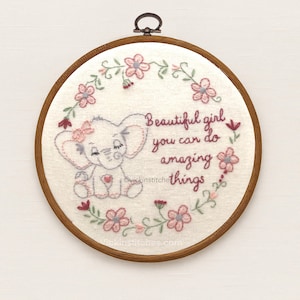 May include: Embroidered hoop art featuring a cute elephant with a pink bow and heart. The text reads "Beautiful girl you can do amazing things" surrounded by a floral wreath. The hoop is framed in a wooden ring.