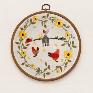 May include: A circular wooden embroidery hoop containing a white fabric panel with a hand-stitched design. The embroidery features a wreath of yellow flowers and green leaves surrounding a farm scene with two chickens, a coop, and a sun.