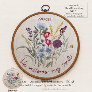 May include: A hand-embroidered floral design in a bamboo hoop. The embroidery features a variety of colorful flowers, including blue, purple, and red blooms, with the text "He restores my soul" stitched in a dark red thread. The text "PSALM 233" is also visible.