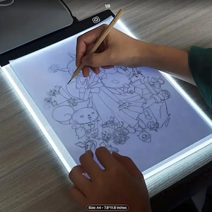 May include: A person is drawing on a lit A4-sized light box (7.8" x 11.8") with a pencil. The drawing is a detailed cartoon with multiple characters. The light box has a black frame and a bright white light.