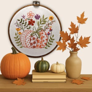 May include: An embroidered pumpkin design in a wooden hoop, surrounded by colorful flowers and leaves. A large orange pumpkin, a green pumpkin, and a small white pumpkin sit on a wooden surface. A vase holds fall leaves.