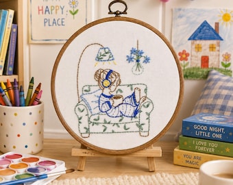 Puede incluir: Un bastidor de bordado circular de madera con tela blanca que presenta un diseño cosido a mano de una persona sentada en un sofá estampado, usando auriculares y sosteniendo una taza. Una lámpara de pie y un jarrón colgante están bordados.
