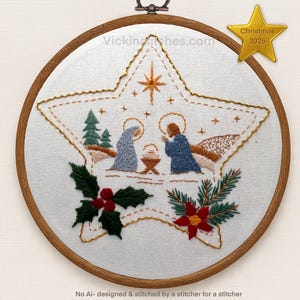 May include: Embroidered Christmas star with a nativity scene, featuring Mary, Joseph, and baby Jesus. The design includes a star, trees, holly, and a poinsettia. The text "Christmas 2025" is on a gold star. The embroidery is set in a wooden hoop.