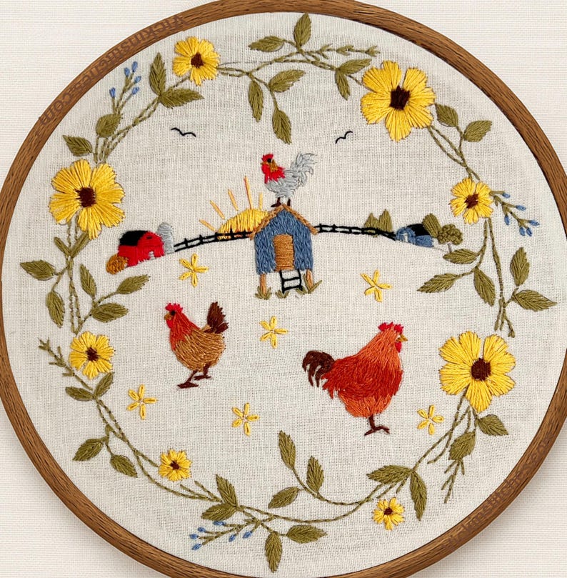 May include: A circular embroidery hoop containing a light-colored fabric with a hand-stitched scene of a farm. The design features a central blue coop with a rooster on top, two chickens, a red barn, and a sunrise, all framed by yellow flowers.