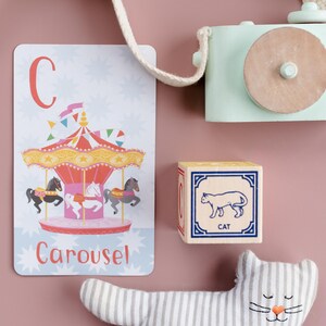 May include: A colorful alphabet flashcard with the letter C and the word "Carousel" printed on it. The card features an illustration of a carousel with horses. A wooden block with the letter C and a picture of a cat is also shown. A striped stuffed cat toy is on a pink background.