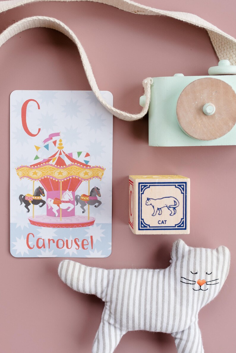 May include: A colorful alphabet flashcard with the letter C and the word "Carousel" printed on it. The card features an illustration of a carousel with horses. A wooden block with the letter C and a picture of a cat is also shown. A striped stuffed cat toy is on a pink background.