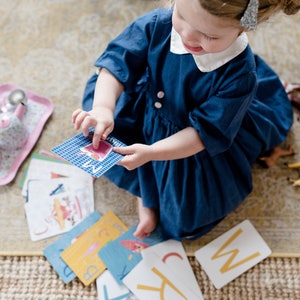 May include: A young child sits on a rug and plays with alphabet flash cards. The cards have colorful illustrations and letters. The child is wearing a blue dress and a white collar. There is a pink tea set and toy horses on the rug.