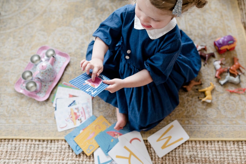May include: A young child sits on a rug and plays with alphabet flash cards. The cards have colorful illustrations and letters. The child is wearing a blue dress and a white collar. There is a pink tea set and toy horses on the rug.