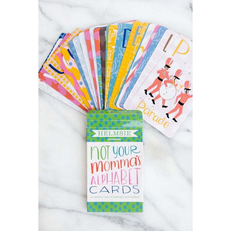 May include: A green box with white polka dots and the text "Helmslie presents Not Your Momma's Alphabet Cards" with a colorful illustration of a parade. The box contains a set of alphabet cards.