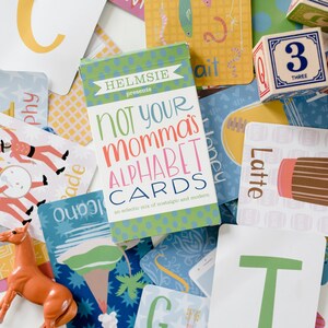 May include: A set of alphabet flash cards with a green and white box that says "Not Your Momma's Alphabet Cards: An eclectic mix of nostalgic and modern". The cards feature illustrations of a parade, a remote, a latte, and a honeybee.