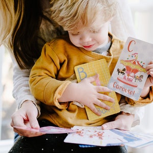 May include: A young child is holding a set of alphabet flash cards. The cards feature colorful illustrations and the letter "C" for "Carousel".
