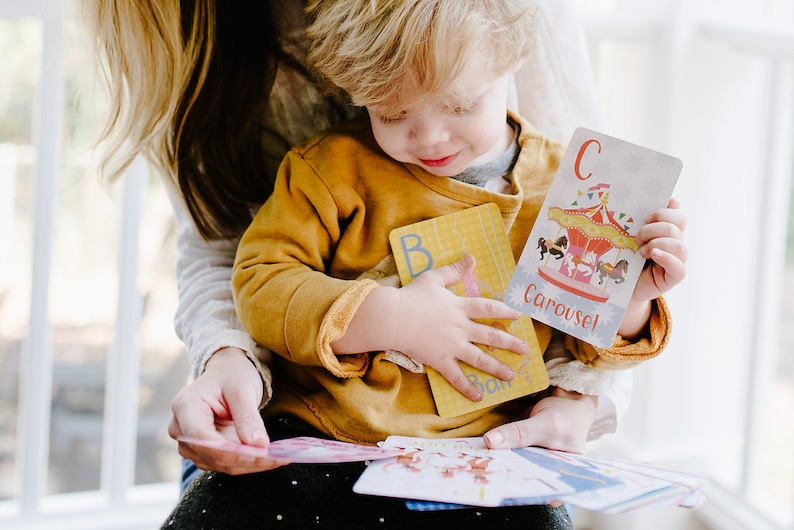 May include: A young child is holding a set of alphabet flash cards. The cards feature colorful illustrations and the letter "C" for "Carousel".