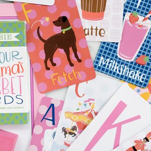 May include: A set of alphabet flash cards with colorful illustrations and playful text. The cards feature a dog, a milkshake, a carousel, a yoga pose, and a ballerina. The box for the cards says "Not Your Momma's Alphabet Cards".