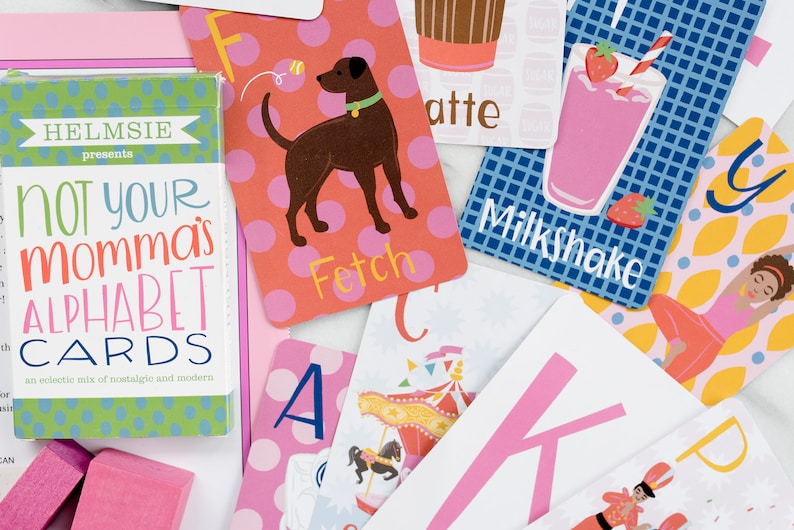 May include: A set of alphabet flash cards with colorful illustrations and playful text. The cards feature a dog, a milkshake, a carousel, a yoga pose, and a ballerina. The box for the cards says "Not Your Momma's Alphabet Cards".