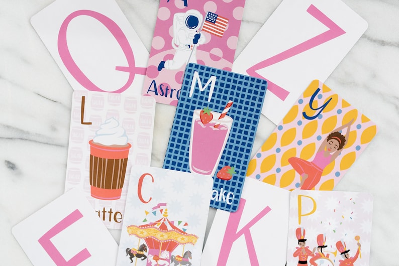 May include: A set of alphabet flash cards with colorful illustrations and pink letters. The cards feature images of a carousel, a milkshake, an astronaut, a yoga pose, a coffee cup, and a marching band.