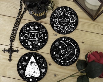 May include: Three circular coasters featuring white graphic designs on a black background. One coaster displays the word OUIJA, another shows a sun face, and the third features a planchette. Each coaster is decorated with stars and symbols.