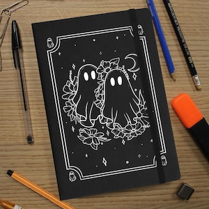May include: Black notebook featuring a white illustration of two ghosts surrounded by flowers and stars. The notebook has a black elastic closure.