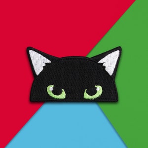 May include: Black felt cat face patch with white ears and green eyes. The patch is on a red, green, and blue background.