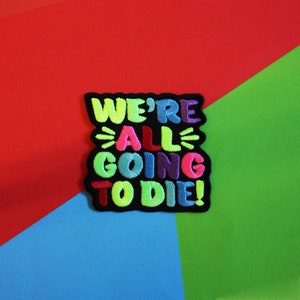 May include: A black embroidered patch with the text "We're All Going To Die!" in colourful letters. The letters are outlined in black and have a 3D effect.