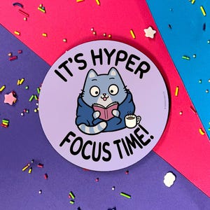 Op de afbeelding: Ronde, paarse sticker met een blauwe kat die een blauwe trui draagt en een boek vasthoudt. De kat zit op een paarse achtergrond met de tekst "It's Hyper Focus Time!" in wit.