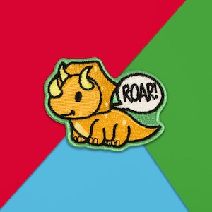 Puede incluir: Parche bordado con un dinosaurio de dibujos animados amarillo y naranja, con contorno verde. El dinosaurio tiene un bocadillo con la palabra "ROAR!" sobre un fondo rojo, verde y azul.