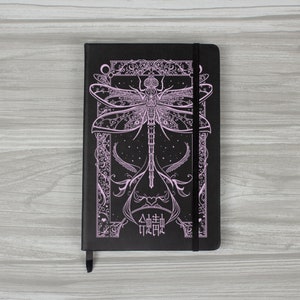 Może przedstawiać: Czarny notatnik z różowym wzorem ważki. Okładka przedstawia ozdobną ilustrację ważki ze skomplikowanymi detalami i dekoracyjnymi obramowaniami. Zawiera czarne zapięcie na gumkę i wstążkową zakładkę.