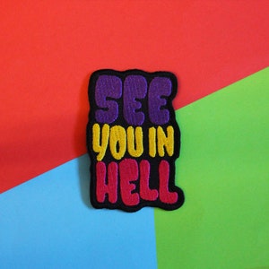 May include: A black embroidered patch with the text "See you in hell" in colourful letters. The letters are purple, yellow, and pink.