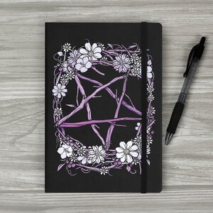 May include: A black notebook featuring a purple pentagram encircled by white flowers and vines. The notebook has an elastic closure and a black pen is positioned beside it.