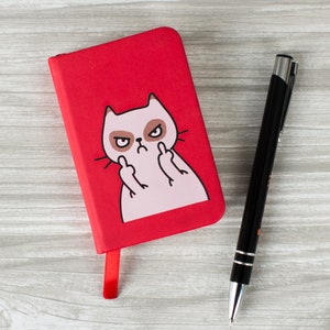 May include: A red notebook featuring a white cat giving the middle finger. The cat has a grumpy expression and is outlined in black. The notebook has a red ribbon bookmark.