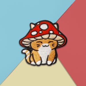 May include: Embroidered patch of a cartoon cat wearing a red mushroom cap with white spots. The cat has orange and white fur, a smiling face, and a striped tail. The patch is set against a background of blue, yellow, and red.