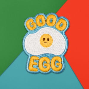 Könnte beinhalten: Gestickter Aufnäher mit einem lächelnden Spiegelei-Design und den Worten "GOOD EGG" in gelben Buchstaben. Das Ei ist weiß mit einem gelben Eigelb und einer blauen Umrandung. Der Aufnäher befindet sich auf einem farbenfrohen Hintergrund.