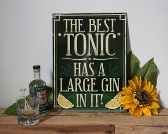 May include: A green metal sign with the text "The Best Tonic Has A Large Gin In It!" with a lemon slice on each side of the text. A bottle of gin and a glass with ice and a lemon wedge are on a wooden surface in front of the sign.