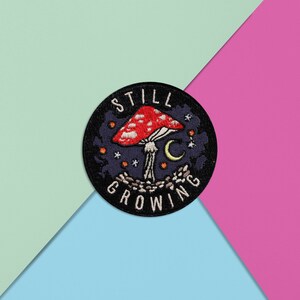 Könnte beinhalten: Schwarzer gestickter Patch mit einem roten und weißen Pilz, einem Halbmond und Sternen. Der Text "STILL GROWING" ist um den Pilz herum geschrieben.