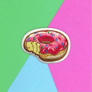 May include: An embroidered patch shaped like a doughnut with a bite taken out of it. The doughnut features a tan dough base, bright pink icing, and small, multi-coloured sprinkles in blue, green, and red, all outlined with a white border.