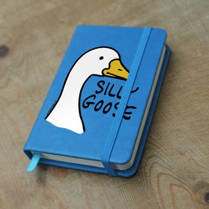 May include: A blue notebook featuring a cartoon goose head and the text "SILLY GOOSE". It has a blue elastic band and bookmark. The notebook is on a wooden surface. The notebook is approximately 14 cm in length.