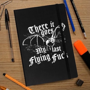 Op de afbeelding: Zwart notitieboek met de tekst "There it goes, my last flying f*ck" in gotisch lettertype, met een vleermuisillustratie. Het notitieboek heeft een zwarte elastische bandsluiting en is omringd door pennen en potloden.